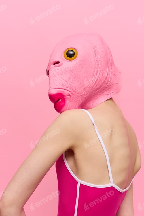 Preview: Skinny woman in a pink Halloween costume with a fish head on her face poses funny against a pink