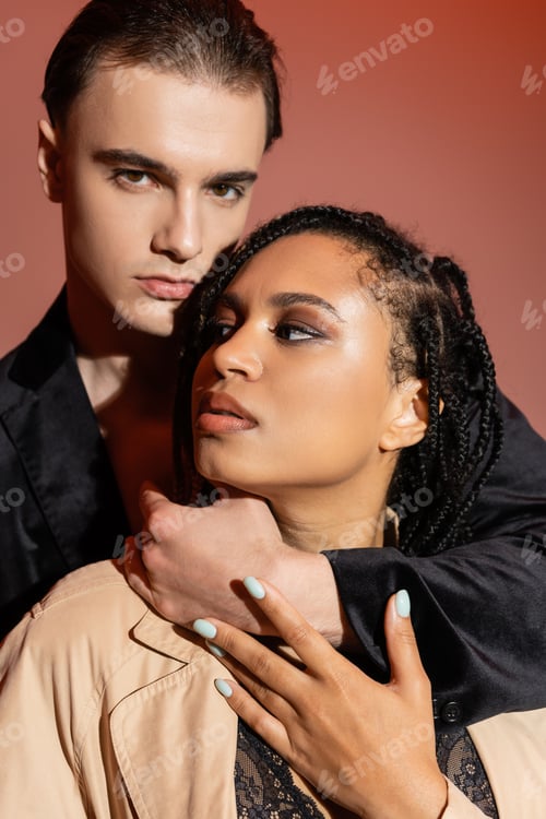 Preview: young charismatic man in black silk blazer looking at camera while hugging neck of sexy african
