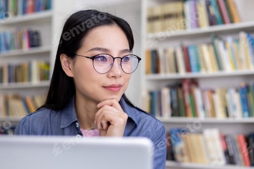 Preview: An Asian woman with glasses contemplates while sitting in a library.