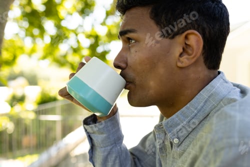 Preview: Thoughtful biracial man drinking coffee and standing on balcony at home