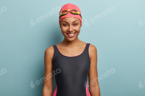 Preview: Happy dark skinned female going to swim, wears pink swimcap and goggles, has sporty body shape, isol