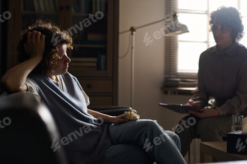 Preview: Sad young brunette female patient of counselor with tissue sitting on couch