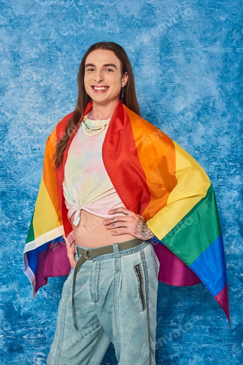Preview: Happy long haired and tattooed gay man in casual clothes with lgbt flag posing and looking at camera