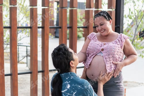 Preview: Hispanic man kneeling and touching pregnant wife's belly outdoors