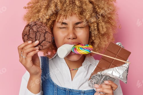 Preview: Indoor shot of positive woman with curly hair enjoys eating delicious sweet treats holds cookie bar