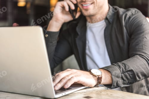 Preview: cropped shot of smiling young freelancer working with laptop and talking by phone in cafe