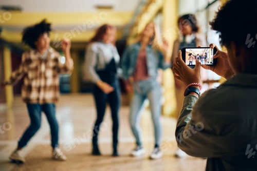 Preview: Close up of teenager using cell phone while photographing his friends at high school hallway.