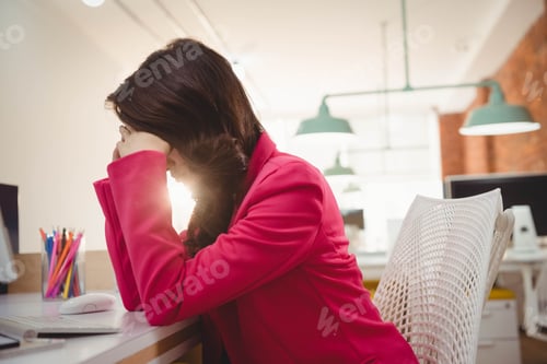 Preview: Asian woman in pink blazer leaning forward covering face amid colored pencils in office, copy space
