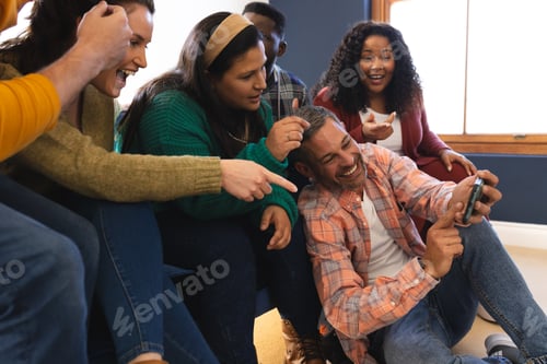 Preview: Happy diverse male and female friends relaxing at home together talking and looking at smartphone