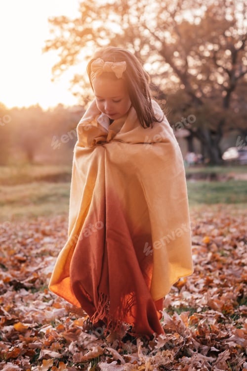 Preview: Little girl 3-4 years old stands in park wrapped in yellow brown stole and looks at foliage in rays