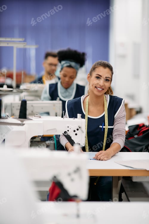 Preview: Happy seamstress sewing at production line in textile factory and looking at camera.