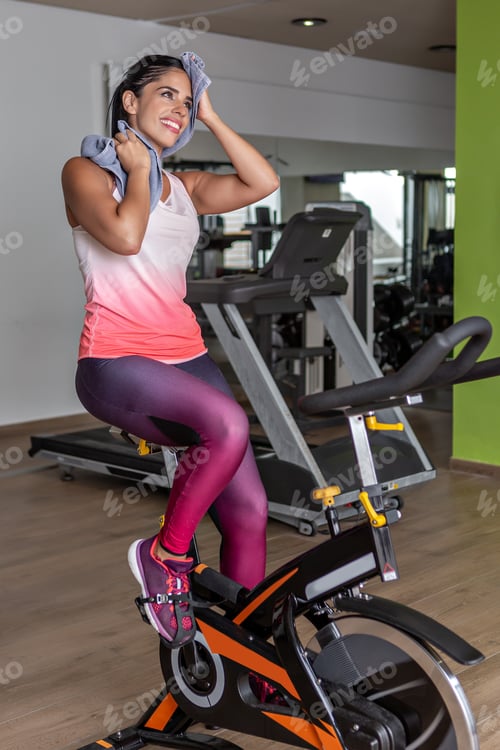 Preview: young sportswoman doing exercise in bike while wiping her face with a towel at gym