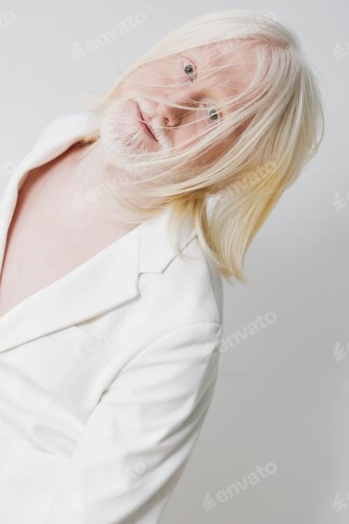 Preview: Portrait of a person with long, light colored hair and a beard, wearing a white blazer against a