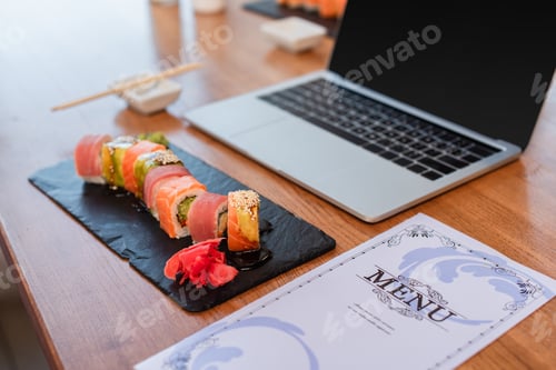 Preview: delicious sushi rolls, menu and blurred laptop with blank screen on table in sushi bar