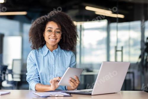 Preview: Portrait of a smiling young African American woman sitting at a desk in the office, holding a