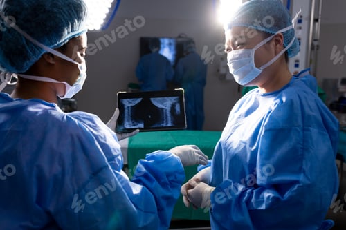 Preview: Diverse female doctors with face masks looking at x-ray on tablet in hospital operating room