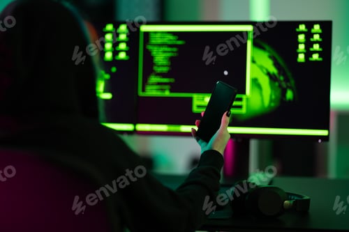 Preview: Hacker holding smartphone in front of dual screens with coding interface