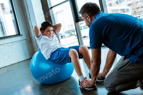 Preview: father helping son sitting on fitness ball and doing sit up exercise at gym