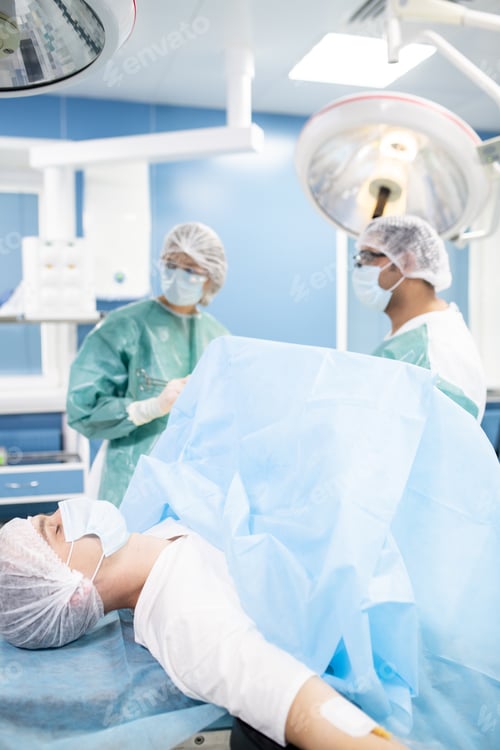 Preview: Young male patient in protective mask and headwear lying against two surgeons
