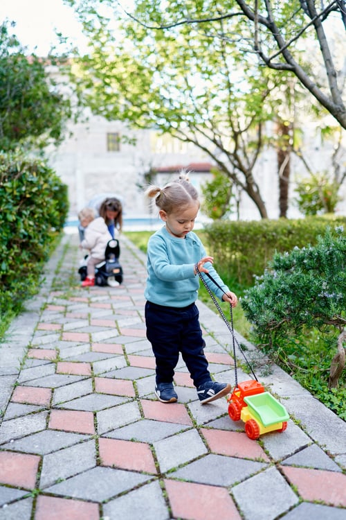 Preview: Little girl with a colorful toy car on a string stands on a paved path in the garden