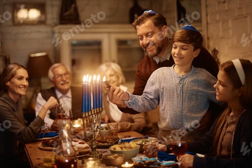 Preview: Happy father and son lighting menorah while celebrating Hanukkah with their family at dining table.