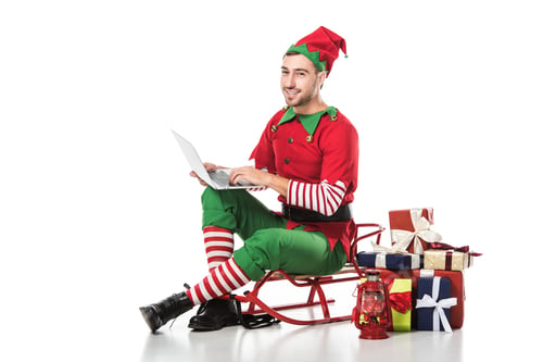 Preview: smiling man in christmas elf costume sitting on sleigh near pile of gifts and using laptop isolated