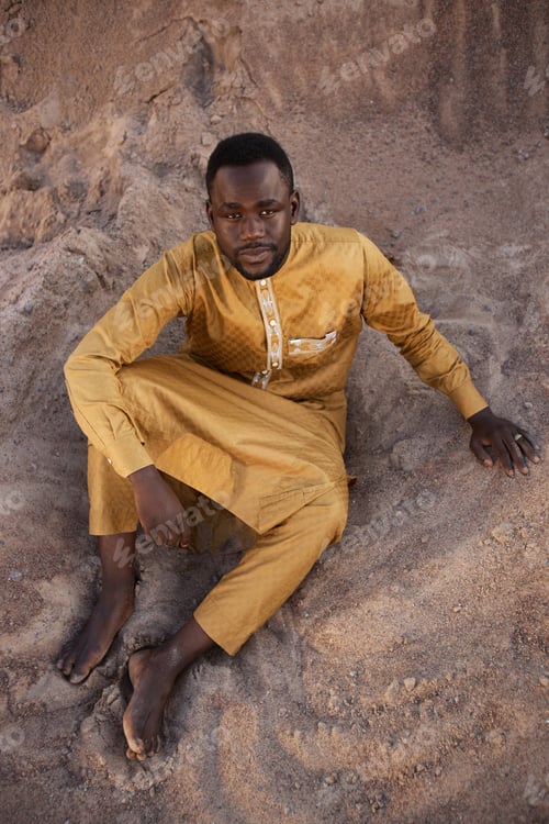 Preview: African American Man in Traditional Kaftan Sitting on Sand in Shadow