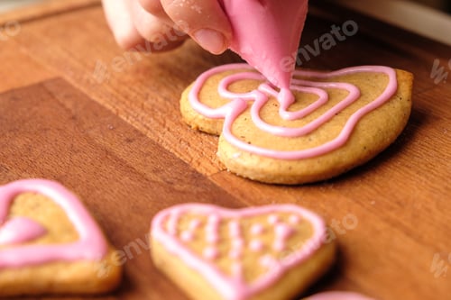 Preview: Icing of Valentines Day. Woman decorating gingerbread cookies in the shape of heart on a wooden