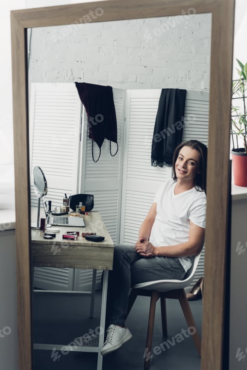 Preview: view through mirror reflection young transgender woman looking at camera at home
