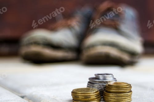 Preview: Stacked coins on blurred background of a pair of dirty old boots. The concept of poverty, homelessne