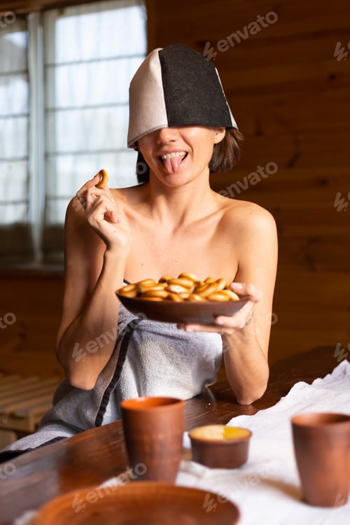 Preview: Young woman in the sauna with a cap on her head sits at a table with round bagels, honey and tea