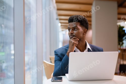 Preview: Portrait of African businessman using laptop computer in coffee shop while looking through window