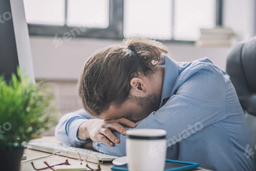 Preview: Young bearded man in blue shirt having a nap at working place