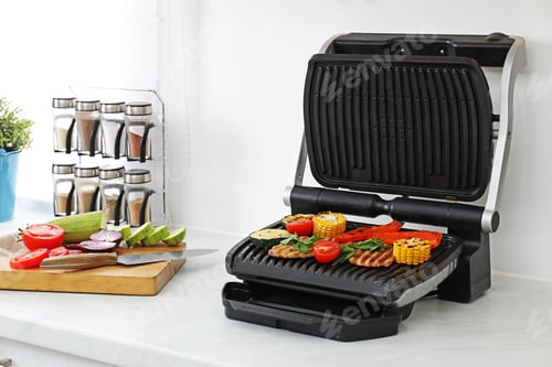 Preview: Electric grill with tasty meat and vegetables on white marble table in kitchen