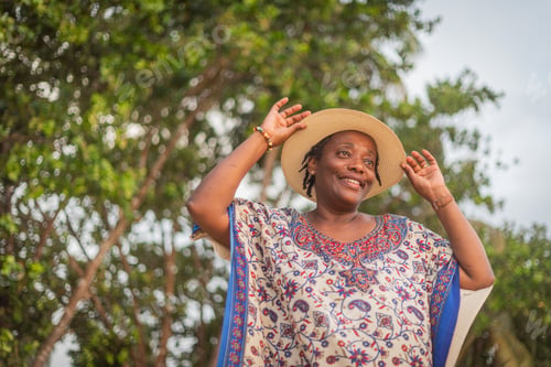 Preview: Happy African woman holding her summer hat and smiling while relaxing outdoors