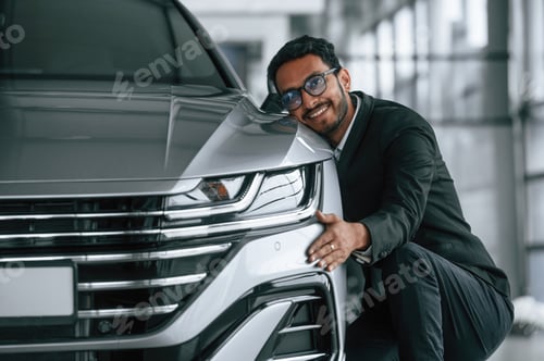 Preview: Embracing the transport. Handsome indian man in suit is in the car showroom with the vehicle indoors