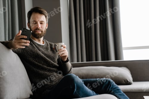 Preview: Bearded man using mobile phone and drinking coffee while sitting on sofa