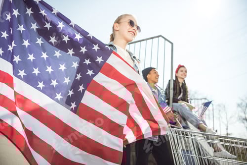 Preview: Blonde young girl with american flag standing near friends having fun at skateboard park