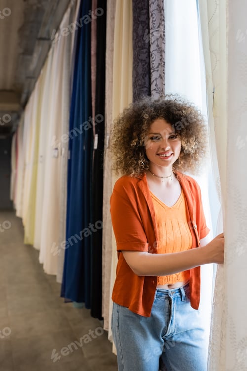 Preview: pretty and curly woman looking at camera near various curtains in textile shop