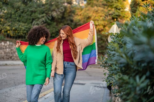 Preview: two gay multiracial lesbian women with rainbow flag gay pride day through the city streets