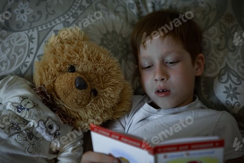 Preview: Child 6-7 years old red-haired boy lies in bed reading a book before going to bed with a teddy bear