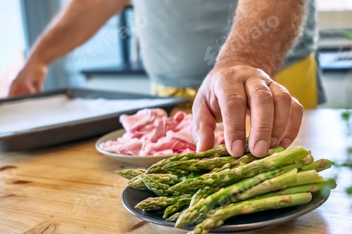 Preview: Man preparing young green asparagus sprouts wrapped in bacon on wooden table in the kitchen.