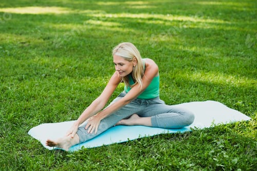 Preview: Woman in sporty outfit stretching her leg on fitness mat during yoga class in park outdoors.