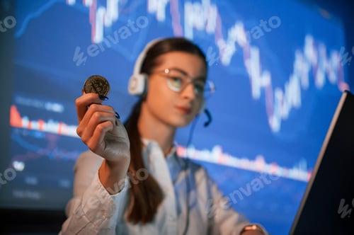 Preview: Holding bitcoin in hand. Female stock broker is working indoors. Big wide display behind her