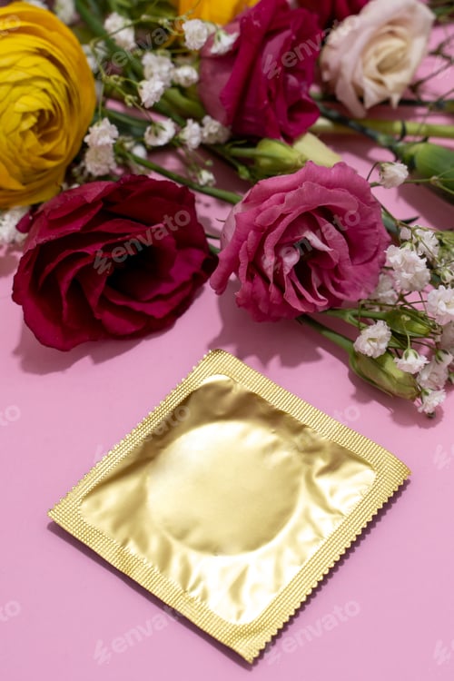 Preview: Golden wrapped condom alongside a delicate flowers on pink background