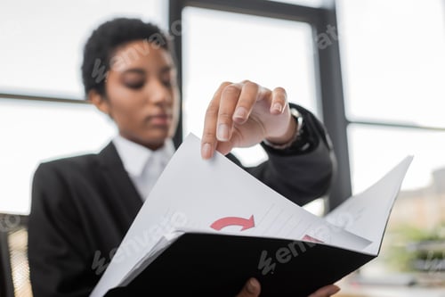Preview: blurred african american businesswoman holding folder with infographics while working in office