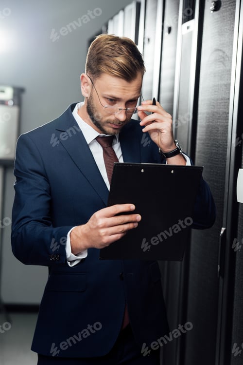 Preview: handsome businessman touching glasses while looking at clipboard