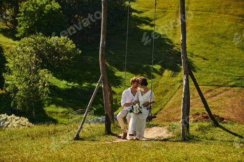 Preview: happy moment of just married multiethnic couple in boho style attire on swing in scenic countryside
