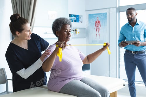 Preview: Diverse senior female patient exercising with band and female doctor advising in hospital room