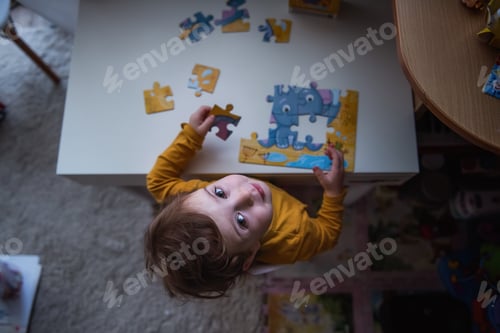 Preview: Little girl in mustard pajamas sits at the table collects puzzle. Happy Child smiles that the puzzle
